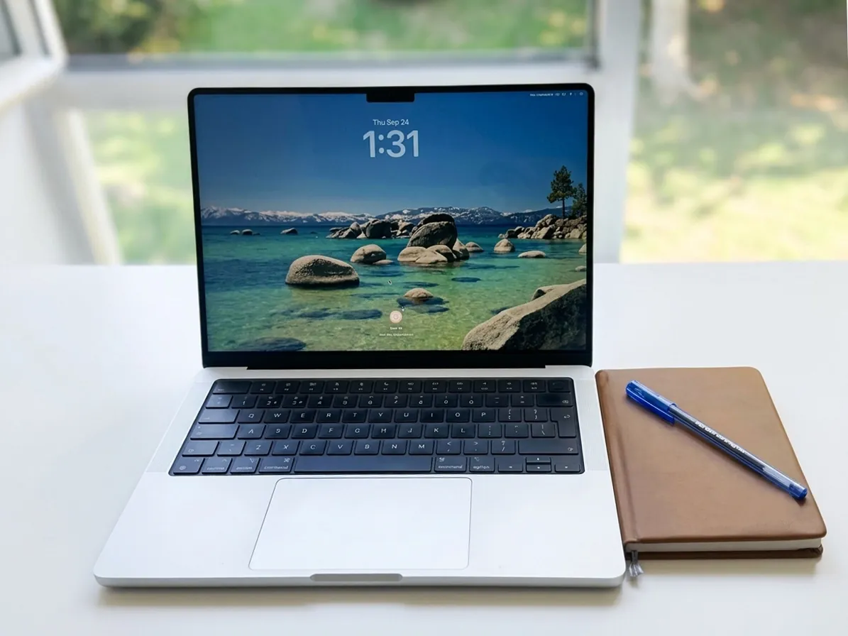 Samama Tahir's web development workspace: an open laptop and a leather notebook on a bright desk